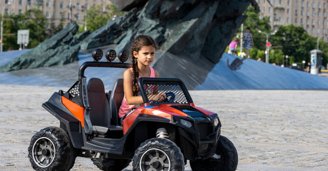 A young girl driving an orange and black ride-on UTV across a paved plaza near a large outdoor monument.