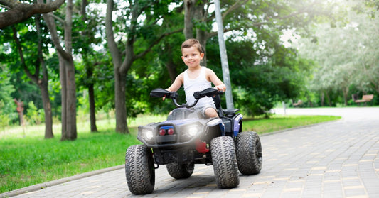 A young boy riding a four-wheeled toy ATV along a paved park path surrounded by green grass and leafy trees.