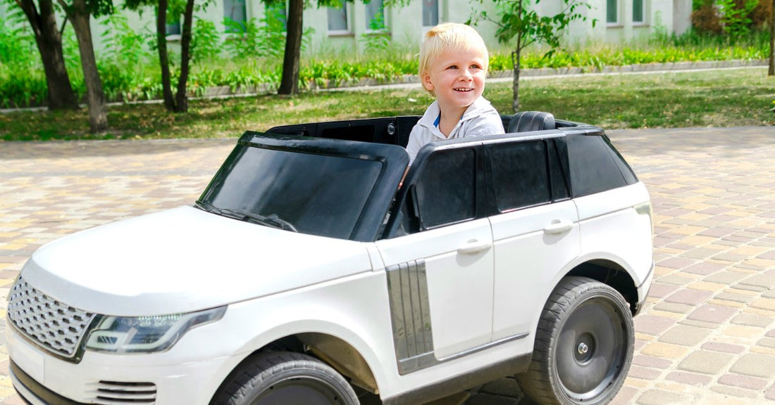 A smiling young boy sitting in a white ride-on SUV on a paved path with trees and greenery in the background.