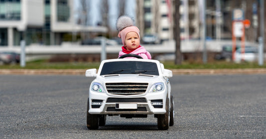 A toddler wearing a pink hat and jacket while driving a white ride-on SUV on a paved road in an urban area.