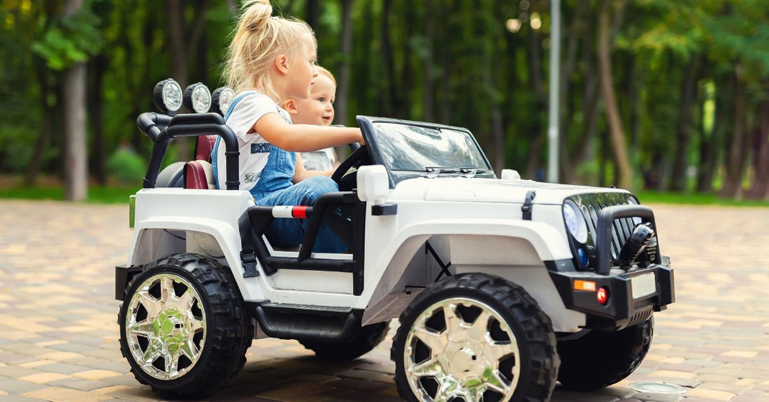 Two small blond children sitting in an electric ride-on SUV. They are driving over a brick path lined with trees.
