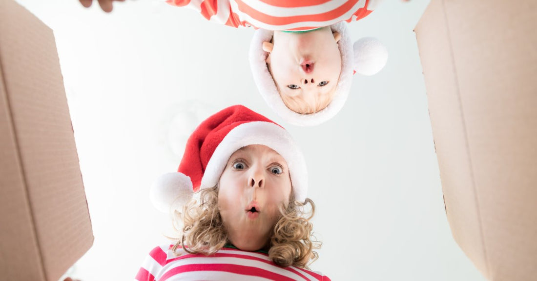 A little boy and girl in red-and-white striped shirts and santa hats lean over an open cardboard box with happily surprised faces.