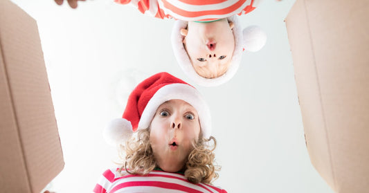 A little boy and girl in red-and-white striped shirts and santa hats lean over an open cardboard box with happily surprised faces.