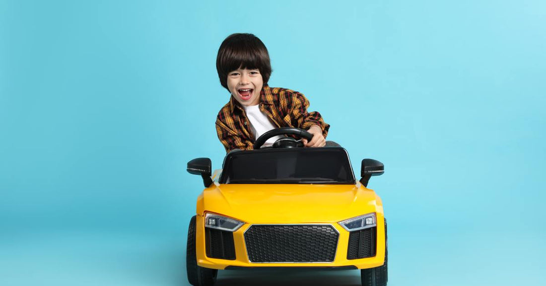 A younger boy wearing a brown plaid shirt smiles and drives a yellow toy car against a light blue background.