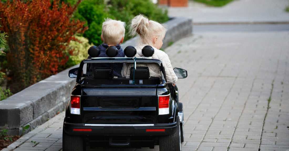 A young boy and girl sitting together in a black two-seat ride-on car, driving along a brick path lined with plants.