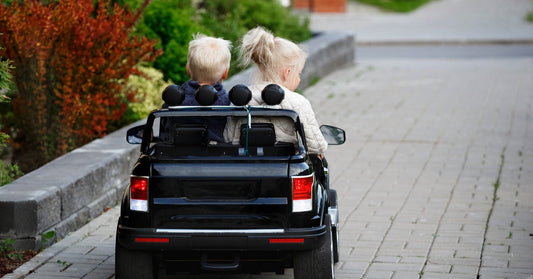 A young boy and girl sitting together in a black two-seat ride-on car, driving along a brick path lined with plants.