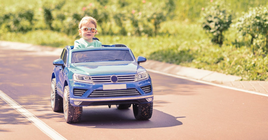 A young girl riding in a children's car across a grass lawn. She has a stuffed animal in the vehicle with her.