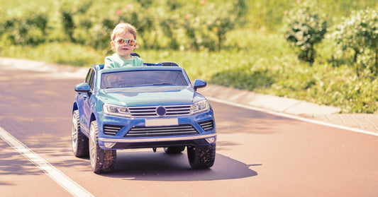 A young girl riding in a children's car across a grass lawn. She has a stuffed animal in the vehicle with her.