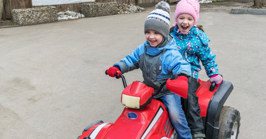 Two small children wearing hats, gloves, and jackets while riding together on a red electric ATV for kids.