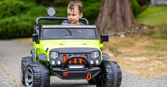 A close-up of a young child driving a green and black electric ride-on car along a path lined with trees.