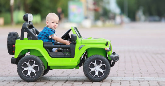 A close-up of a young child sitting in the driver's seat of a green electric ride-on vehicle for kids. The car is parked outside.