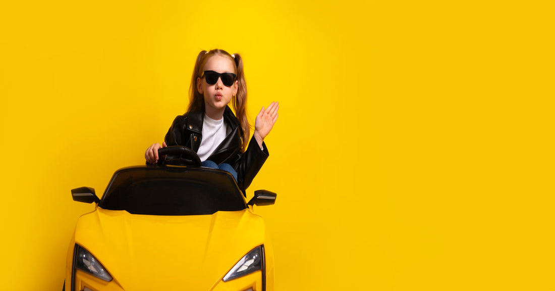 A young girl wearing a black leather jacket and sunglasses sits in a yellow ride-on car. The background is yellow.