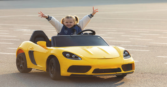 A little girl in a bomber jacket sitting in a bright yellow electric ride-on car. She smiles and holds her arms in the air.