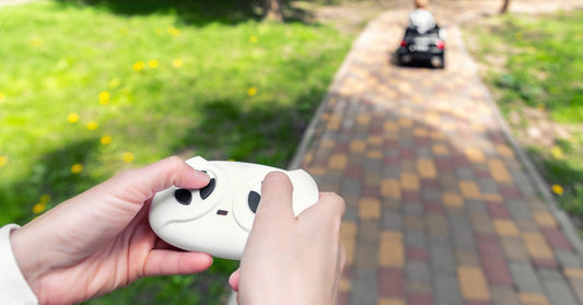 A person's hands operating a remote control, with a child driving a motorized toy car down a path in the background.
