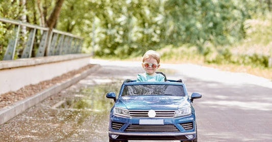 A young boy wears sunglasses as he drives his ride-on car down the sidewalk on a sunny day. He's smirking.