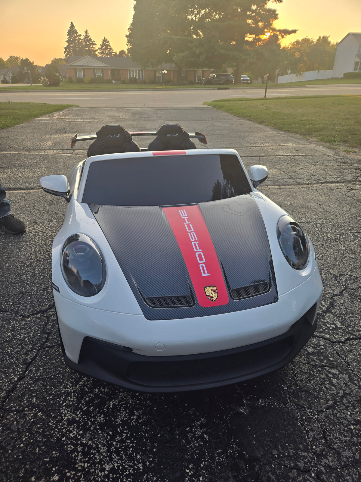 A white Porsche-branded toy car with a black hood and red stripe, photographed outdoors on a driveway at sunset