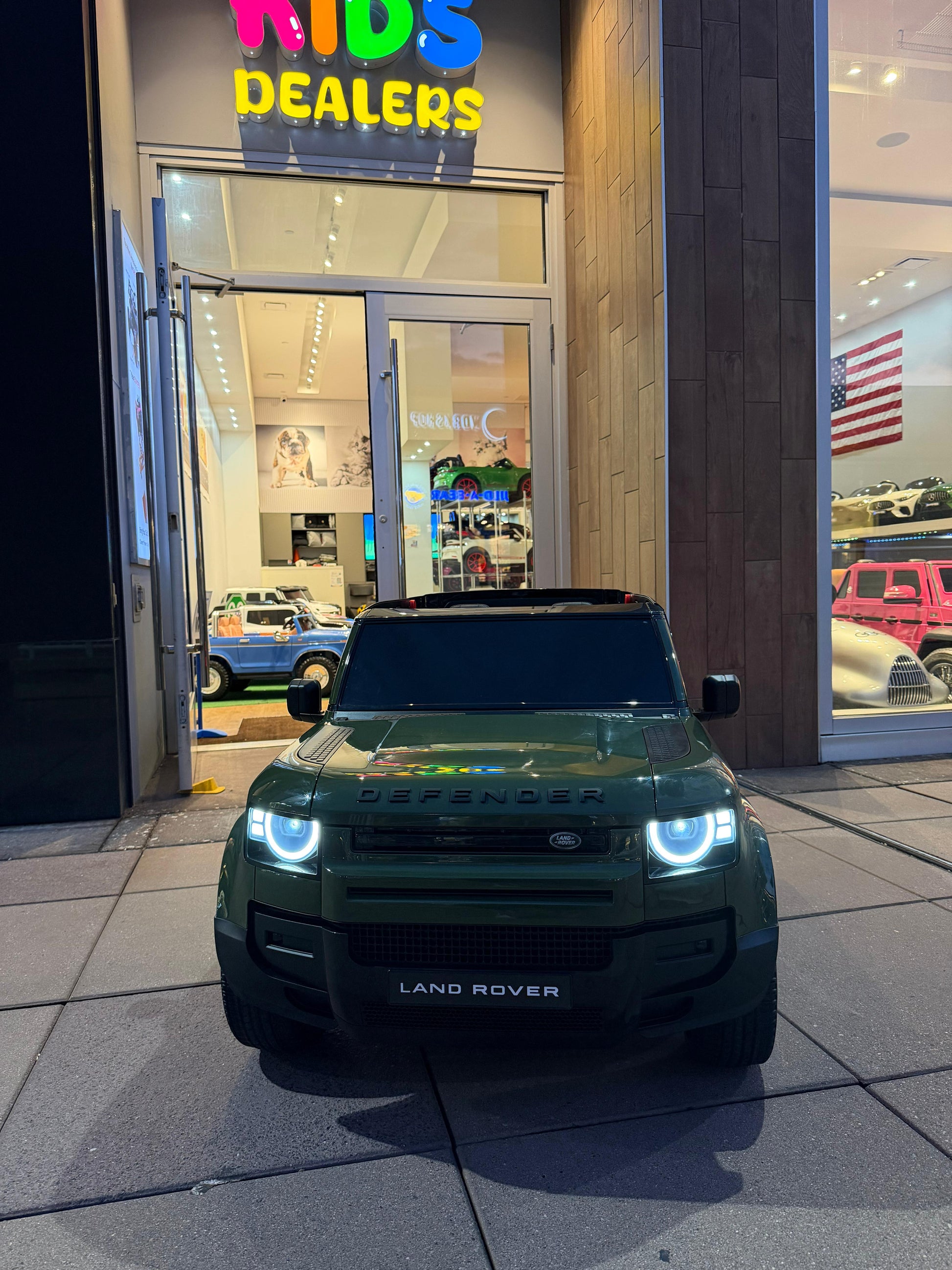 A dark green Land Rover Defender toy car parked in front of a children's toy dealership storefront at night.