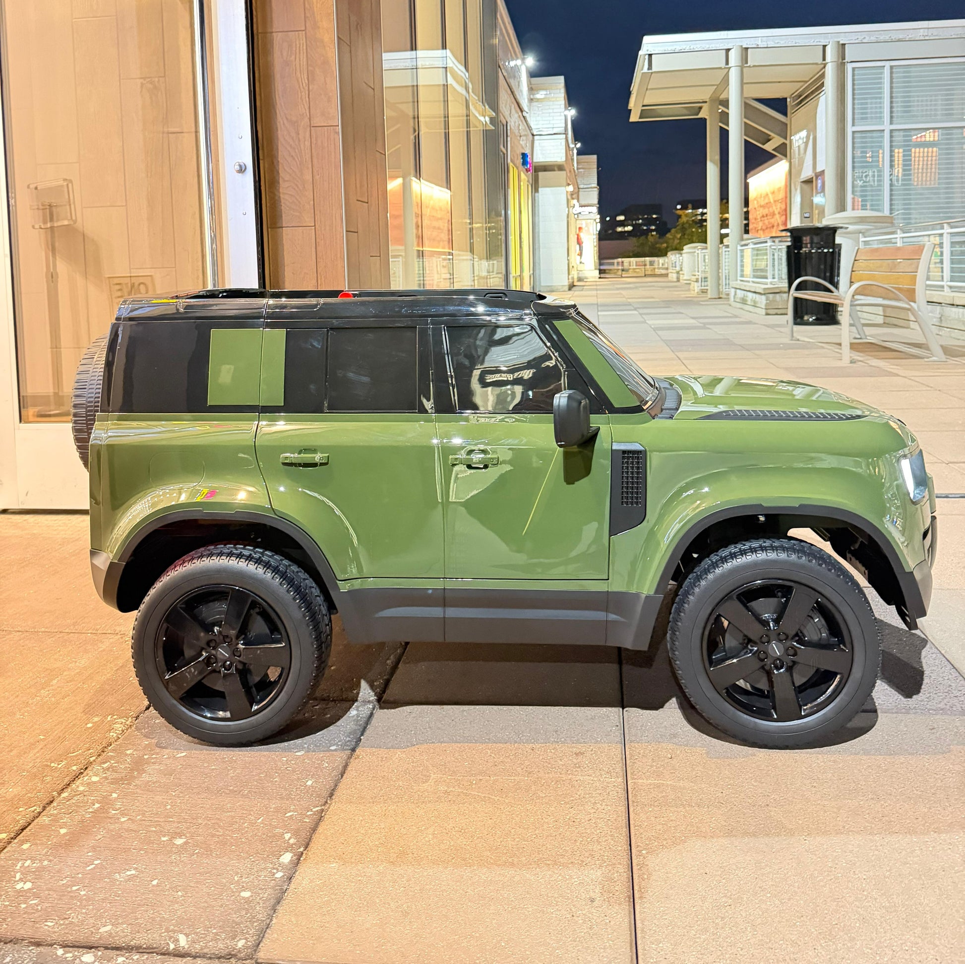 A green defender SUV ride on parked on a paved outdoor walkway at night, with modern glass buildings in the background