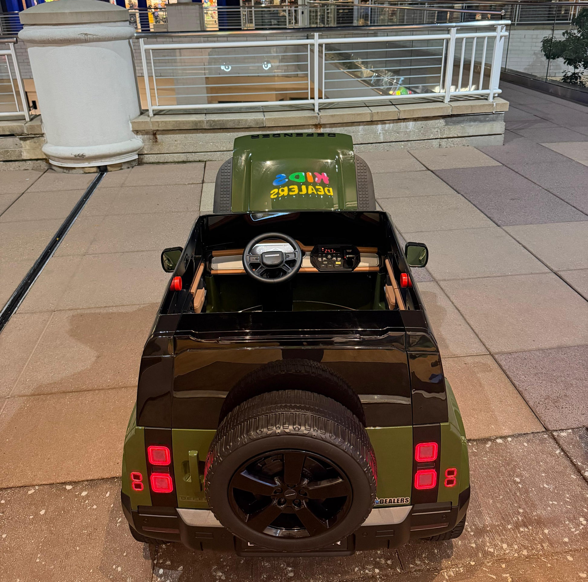 A child's electric ride-on toy car styled like a land rover defender, viewed from the rear. The toy car features an olive green body, a black roof, a black spare tire on the back